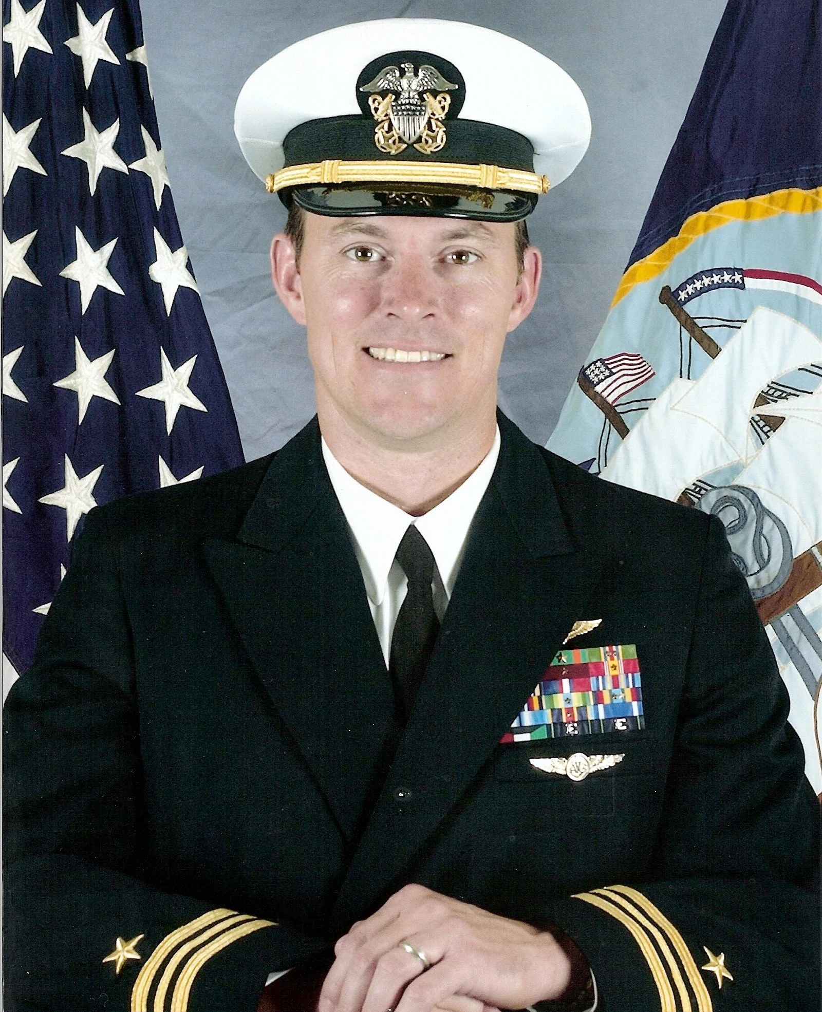 A man in a Navy uniform with a white cap and black jacket, standing in front of American and military flags, smiling.