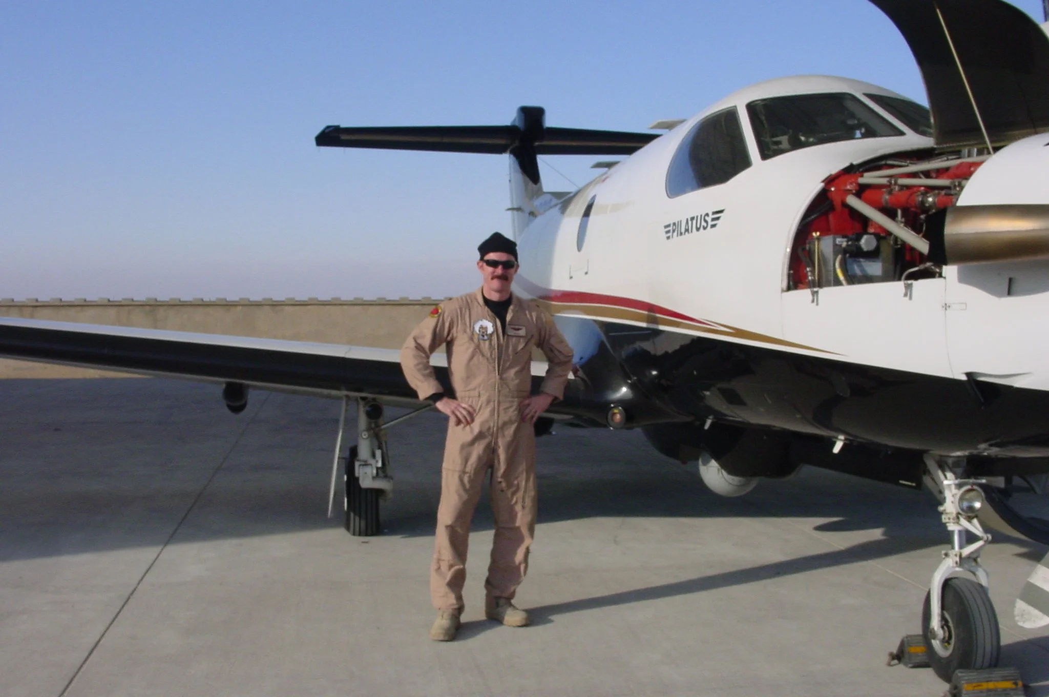 A man in a tan pilot jumpsuit with patches, wearing sunglasses, standing next to a small white aircraft with its engine exposed, on a tarmac.
