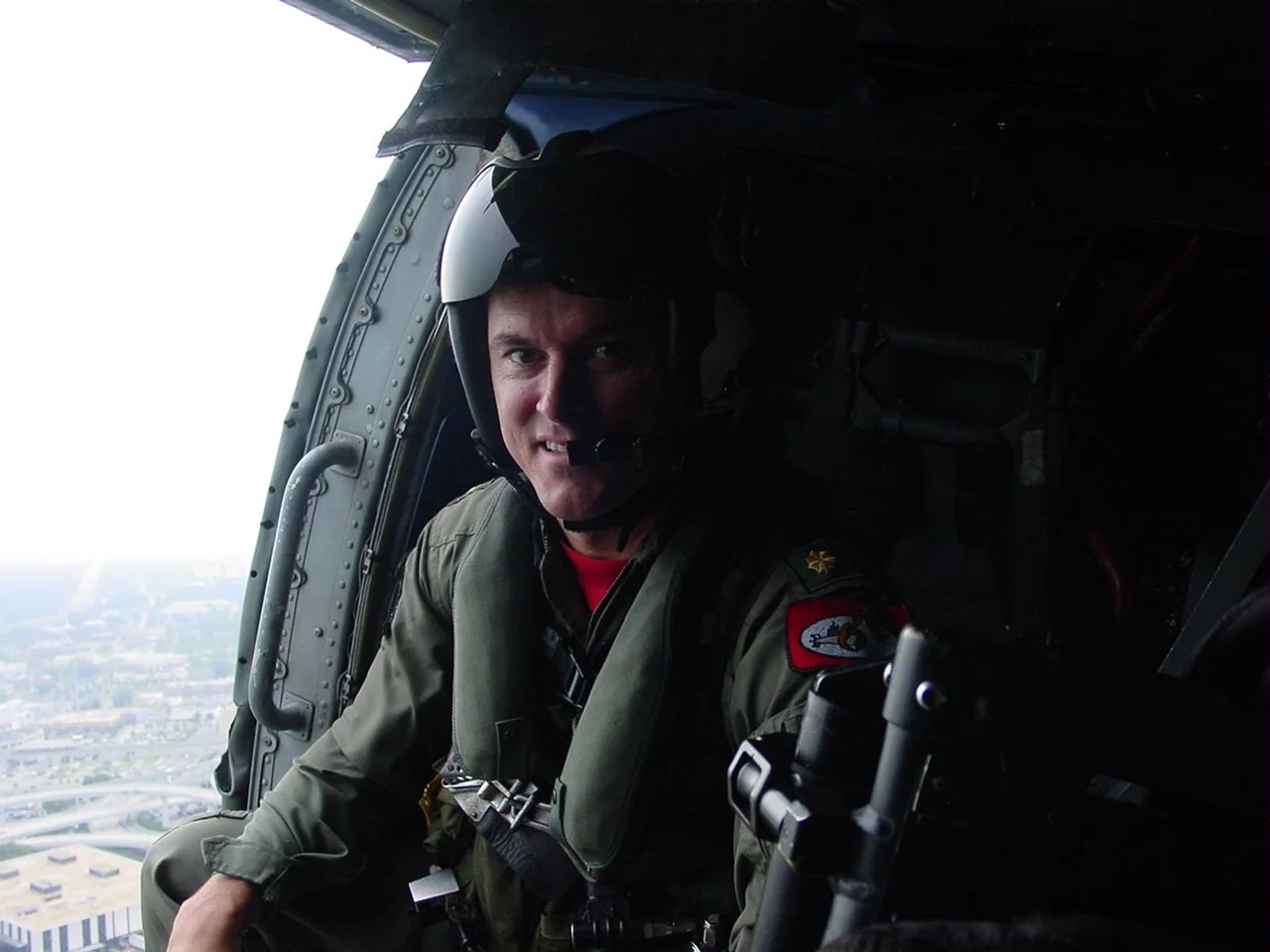 A man in a flight suit and helmet sitting inside the cockpit of an aircraft, with a view of a city and landscape below through the open door.
