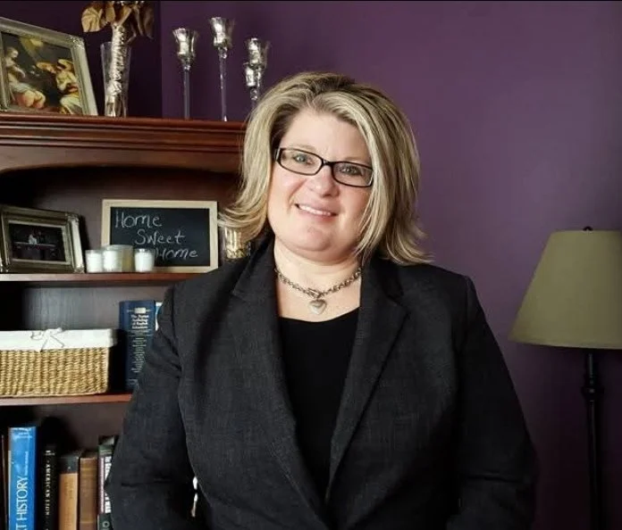 A woman with shoulder-length blonde hair, glasses, and a black blazer stands in a room with purple walls, a lamp, a bookshelf, and a small chalkboard sign that reads "Home Sweet Home."