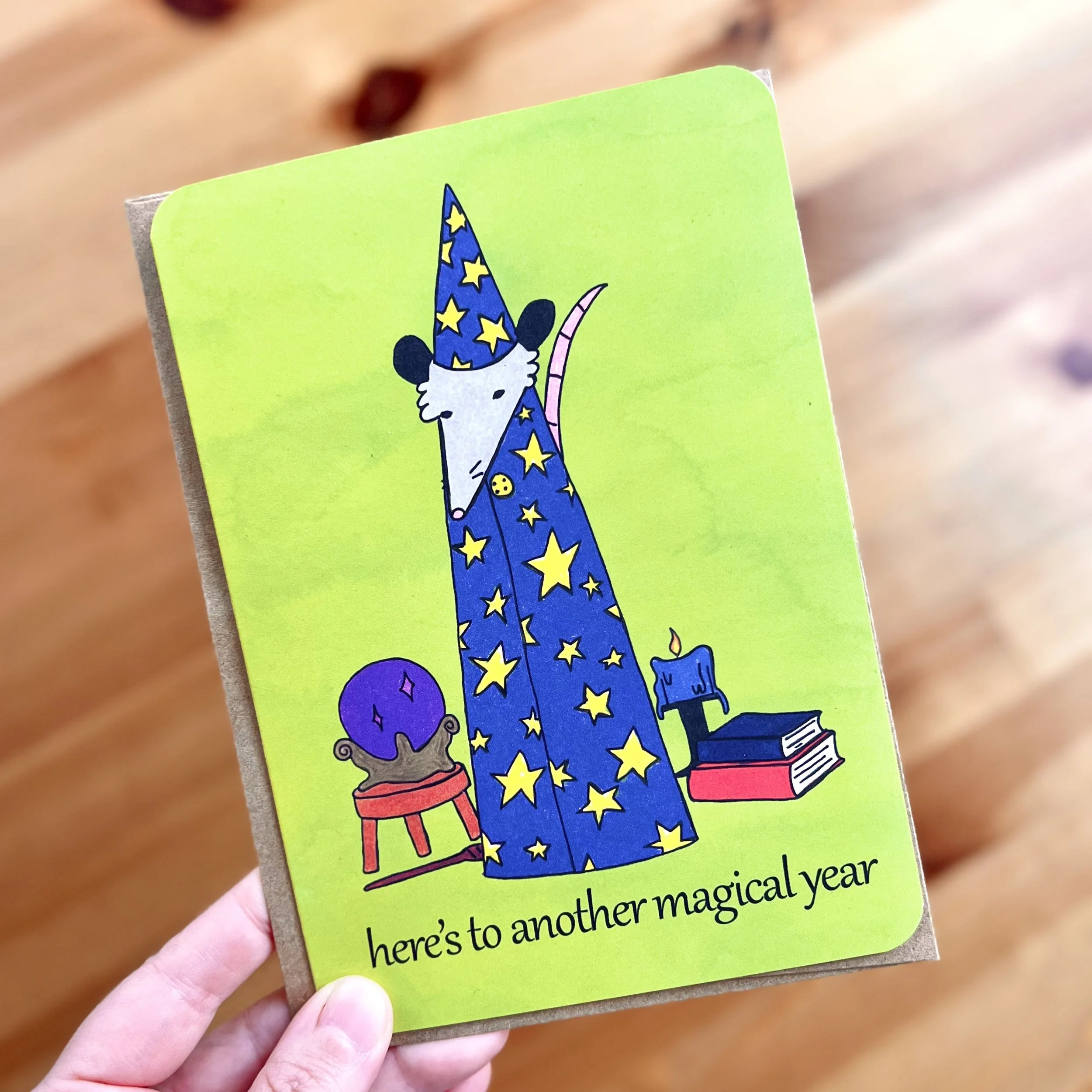 A greeting card with colored pencil drawing of a wizard possum on it. They are wearing a blue cloak with yellow stars all over it with a matching pointy hat. To their right is a stack of two books and a lit candle. To their left is a crystal ball on 