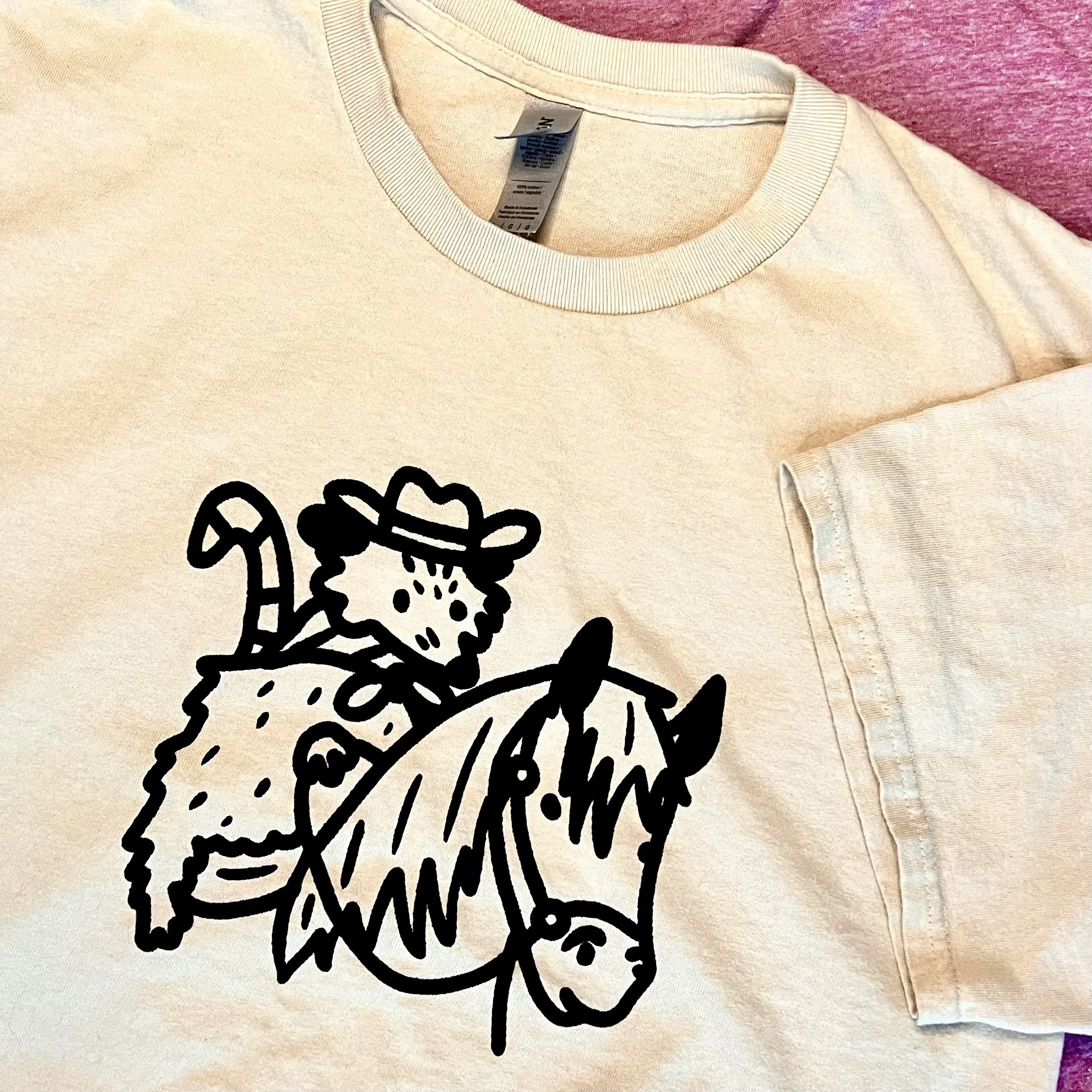 A photo of an off-white t-shirt with a line art illustration of a cowboy possum riding a horse on it.