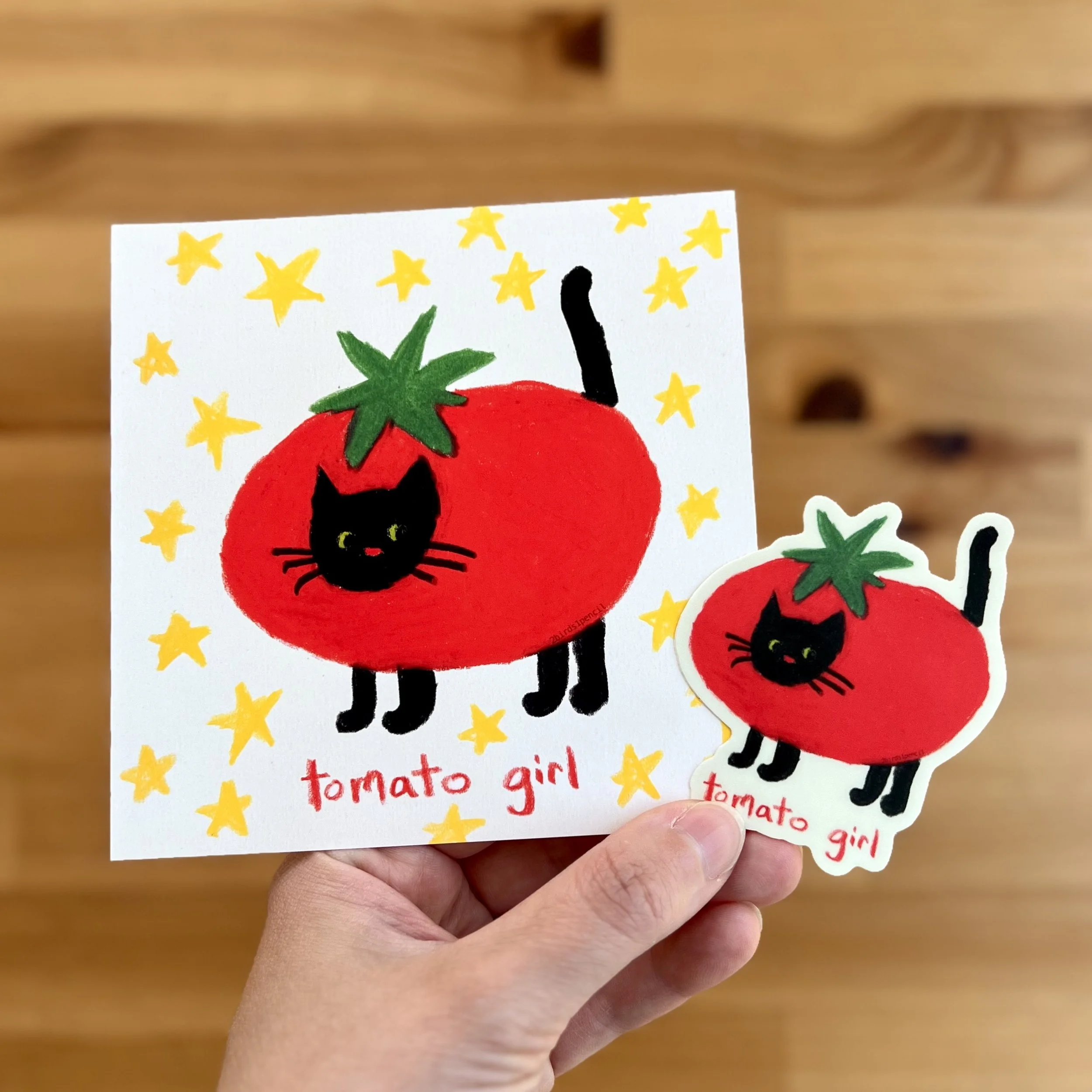 An art print and a sticker of an illustration of a black cat in a tomato costume. They're surrounded by yellow stars and text beneath them reads, "tomato girl".
