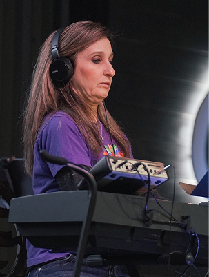 A woman with long brown hair, wearing a purple shirt and headphones, working with audio equipment on a black table, in a dark room with a bright light in the background.