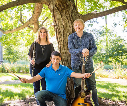 Three musicians outdoors under a large tree, with one seated holding drumsticks, a guitar, and two standing with musical instruments.