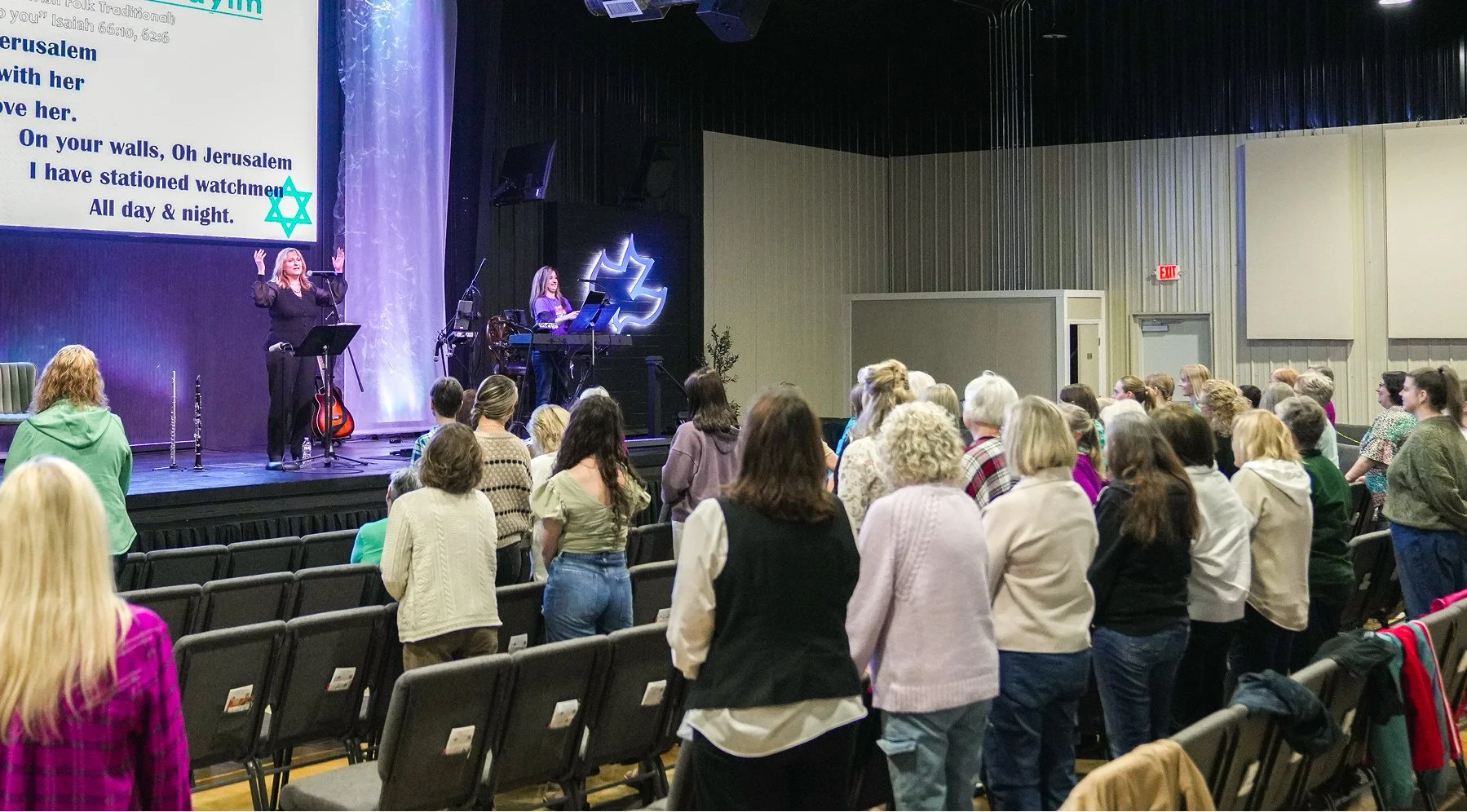 A group of people gathered in an auditorium attending a religious event with a woman singing on stage, along with a person playing keyboard and an audience watching and standing.