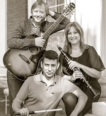 Three people, two women and one man, posing for a photo with musical instruments including an acoustic guitar, a clarinet, and a drumstick, in a house setting.