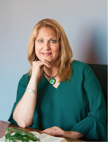 A middle-aged woman with blonde hair sitting at a desk, wearing a green blouse and a necklace with a green stone, resting her chin on her hand and smiling at the camera.