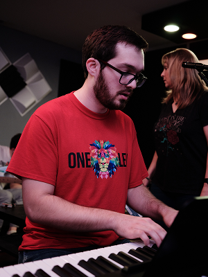 A young man playing the piano in a recording studio, wearing glasses and a red t-shirt with a colorful graphic and text.