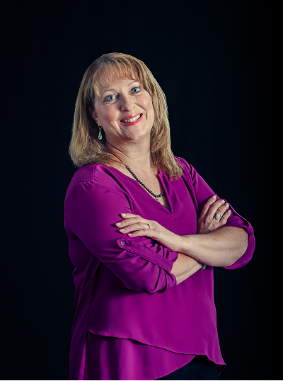 Portrait of a smiling middle-aged woman with blond hair, wearing a purple blouse, crossing her arms, against a dark background.