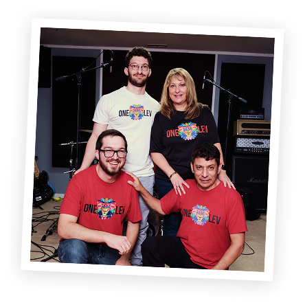 Four people in a music studio, three men and one woman, wearing matching 'One Leh' T-shirts, with musical equipment behind them.