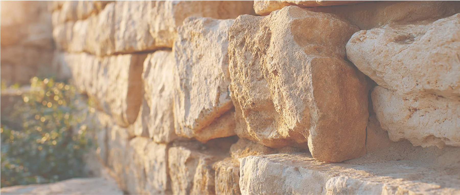 Close-up of a stone wall made of irregularly shaped, beige and light brown stones, with sunlight illuminating the surface.