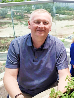 A smiling man in a blue polo shirt sitting at an outdoor table near a body of water with a glass railing behind him.