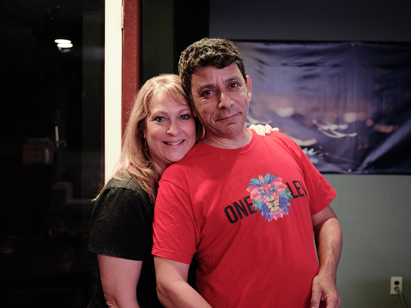 A woman and a man standing close together indoors, smiling at the camera. The woman has blonde hair and is wearing a black shirt, while the man has dark hair and is wearing a red T-shirt with a floral design and the words 'ONE LOVE' on it.