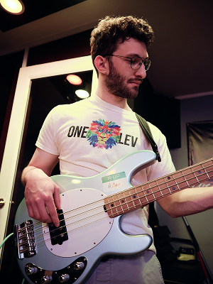 A man with glasses, curly hair, and a beard playing a white electric bass guitar indoors, wearing a white t-shirt with colorful heart-shaped graphic.