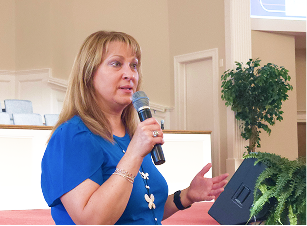 A woman with shoulder-length blonde hair wearing a blue top, speaking into a microphone in an indoor setting with plants and a podium in the background.