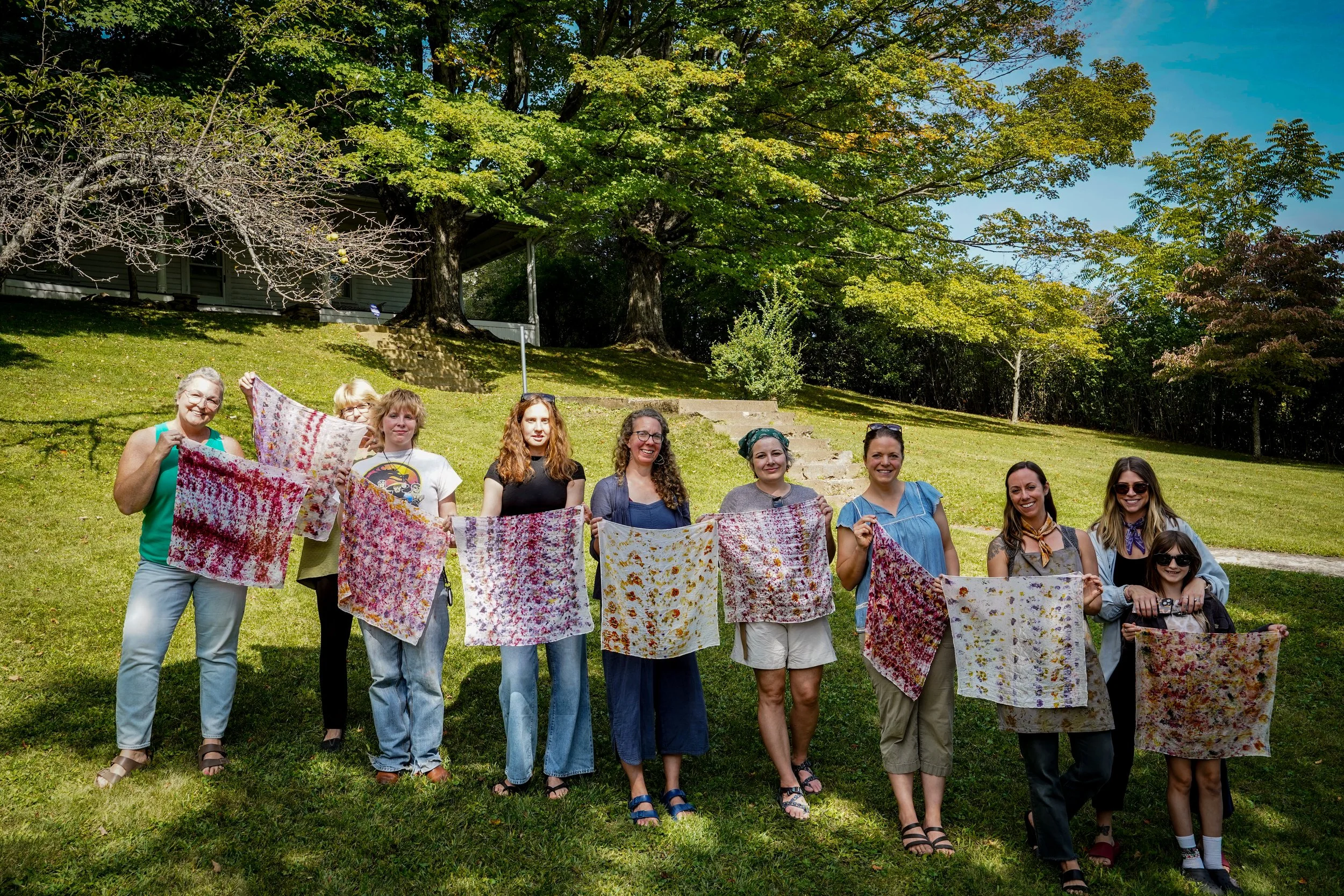 A group of eleven women standing outdoors on a grassy area, holding colorful tie-dye fabric squares. The background features a large tree and a house with stairs leading up a hill. The women are smiling and appear to be participating in a craft or ou