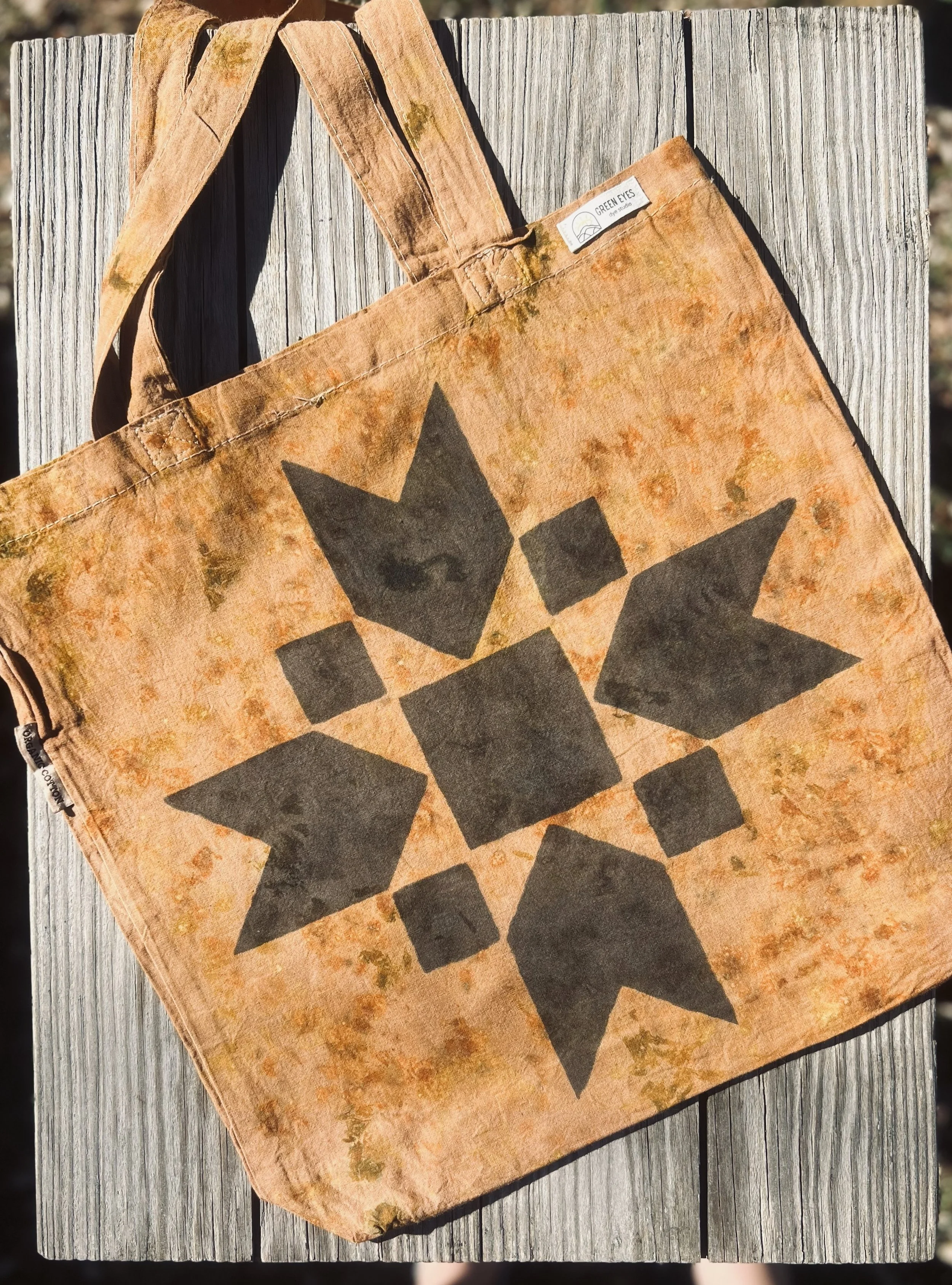 A weathered, brown tote bag with black geometric shapes on the front, resting on a wooden surface outdoors.