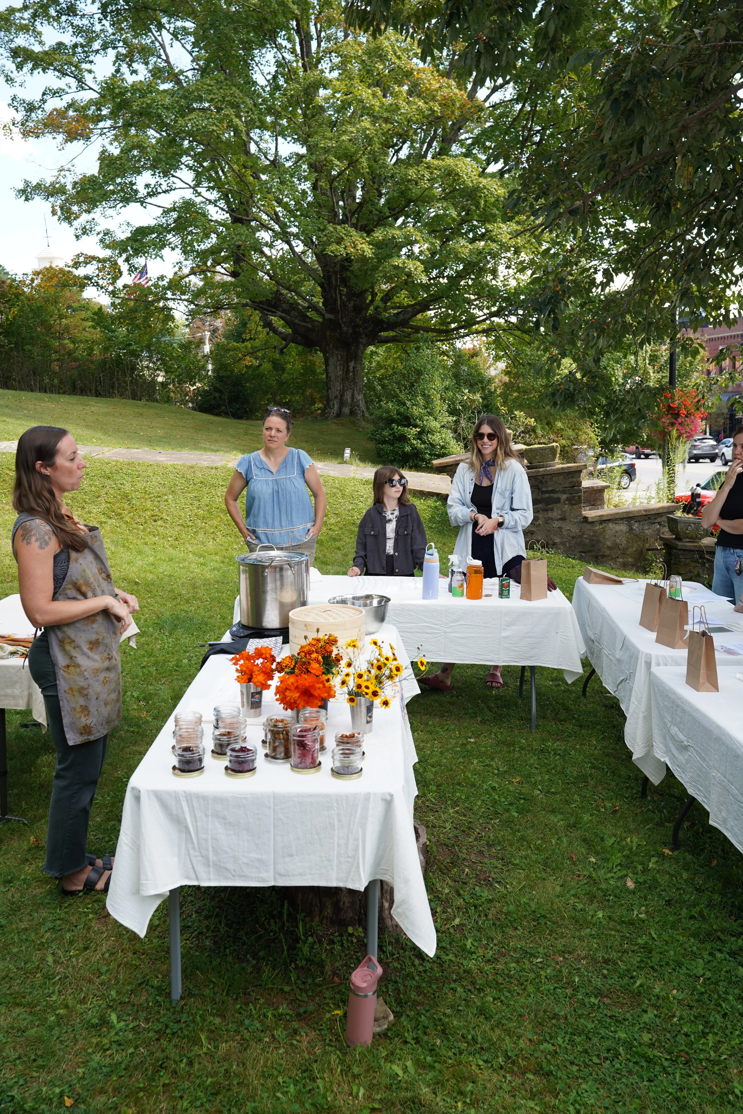 People gathered around outdoor tables with dried plants, leaves, and flowers in a park setting under a large tree.