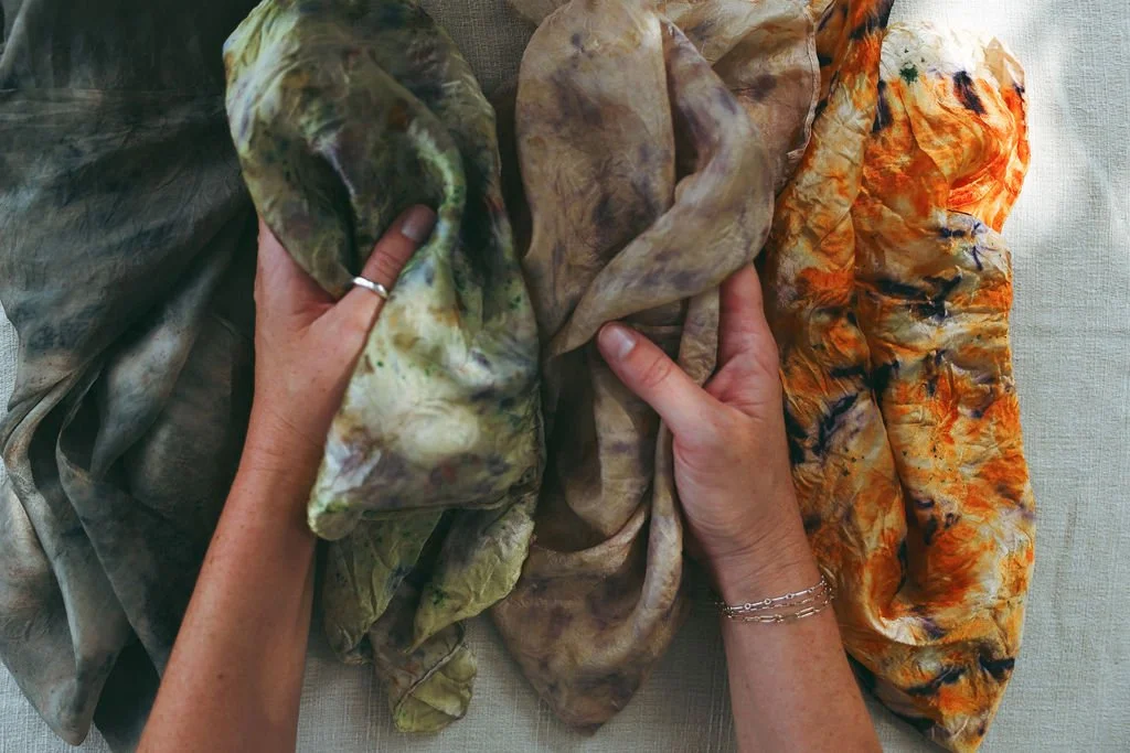 Hands holding scarves with tie-dye patterns in shades of green, brown, and orange laid out on a flat surface.