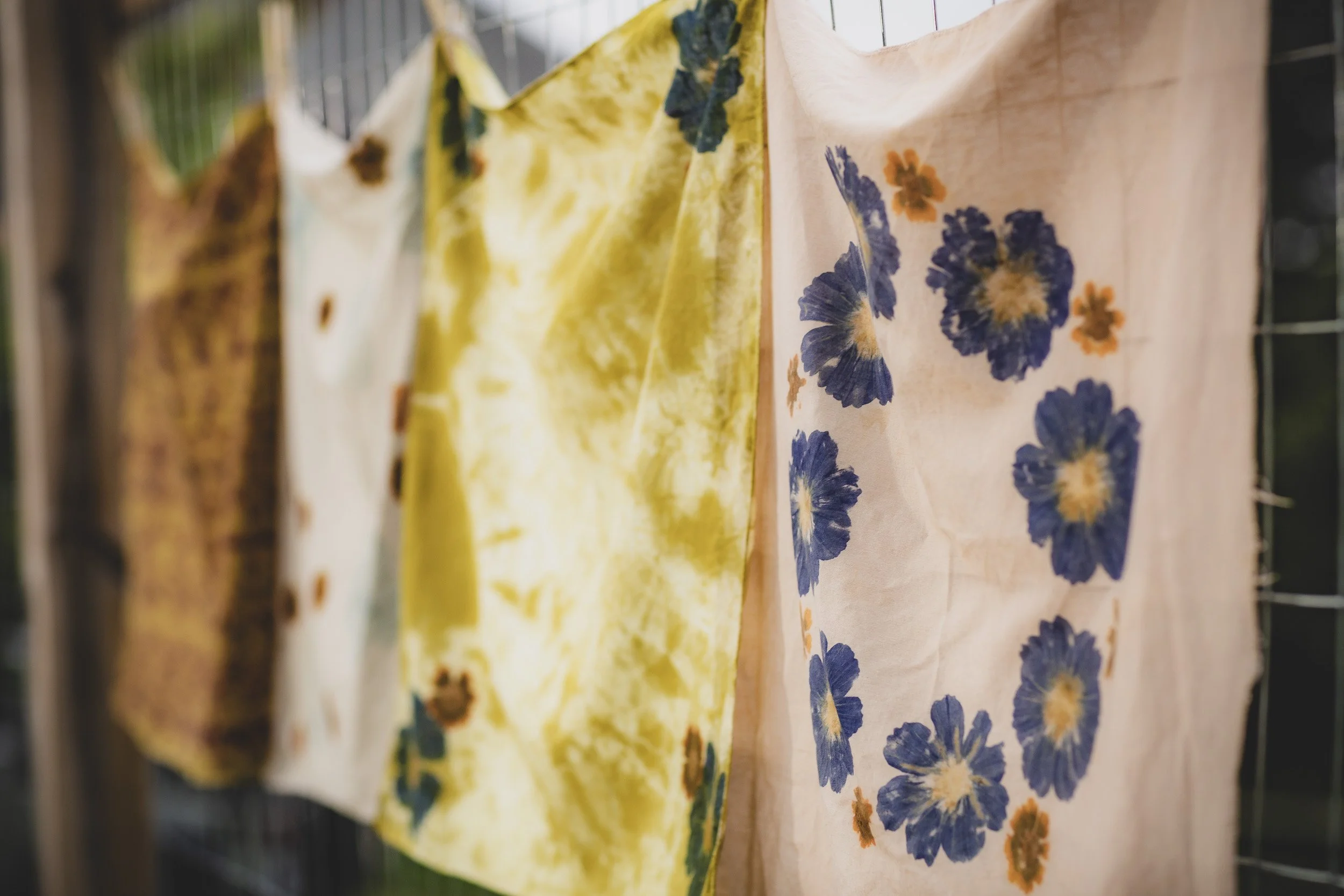 Colorful tie-dye and floral-printed fabric hanging on a wire fence.