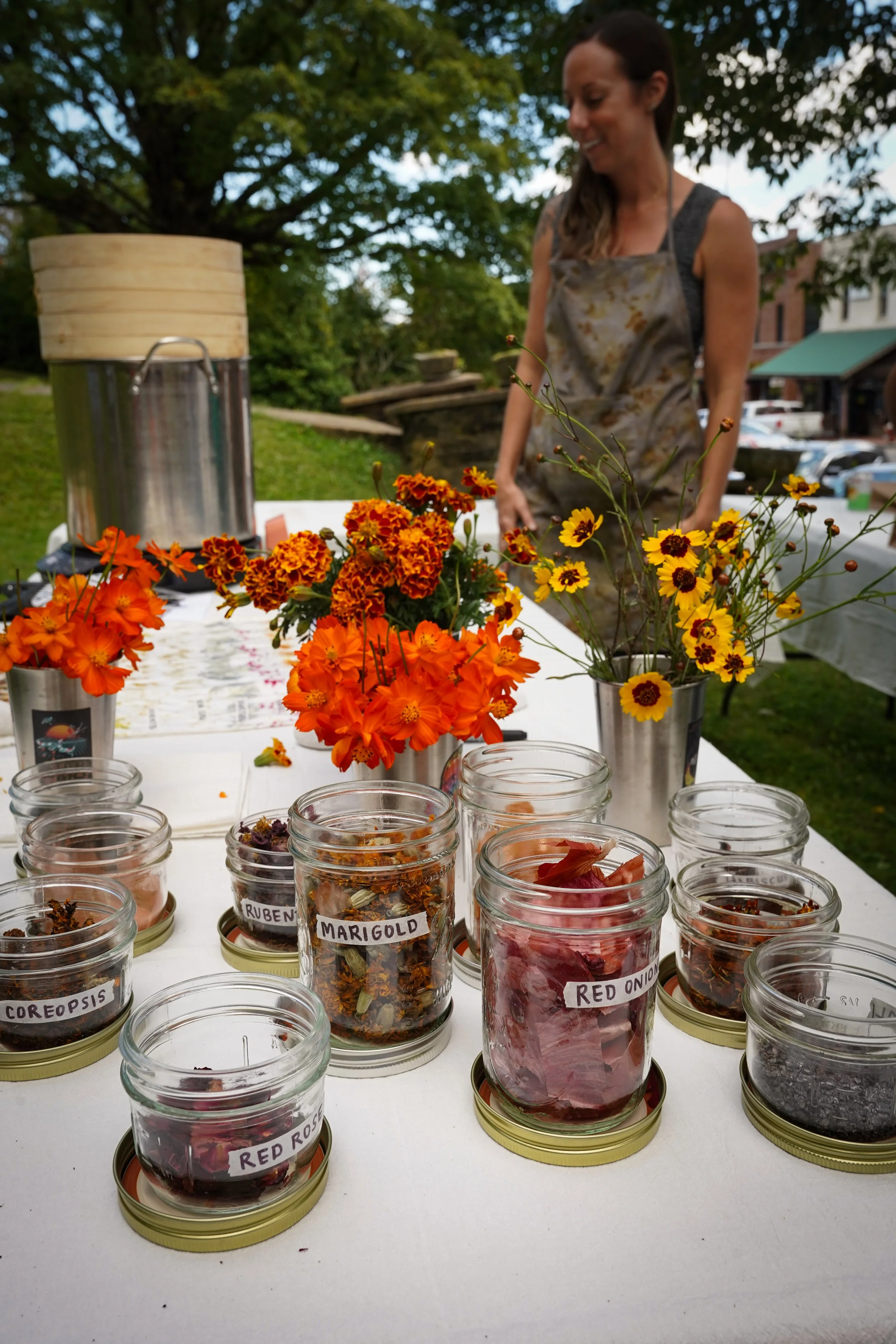 A woman wearing an apron stands behind a table with jars of dried herbs and flowers, labeled with names like 'Coreopsis,' 'Ruben,' 'Marigold,' and 'Red Onion.' The table also has vibrant orange and yellow flowers in metal containers, and a large vint