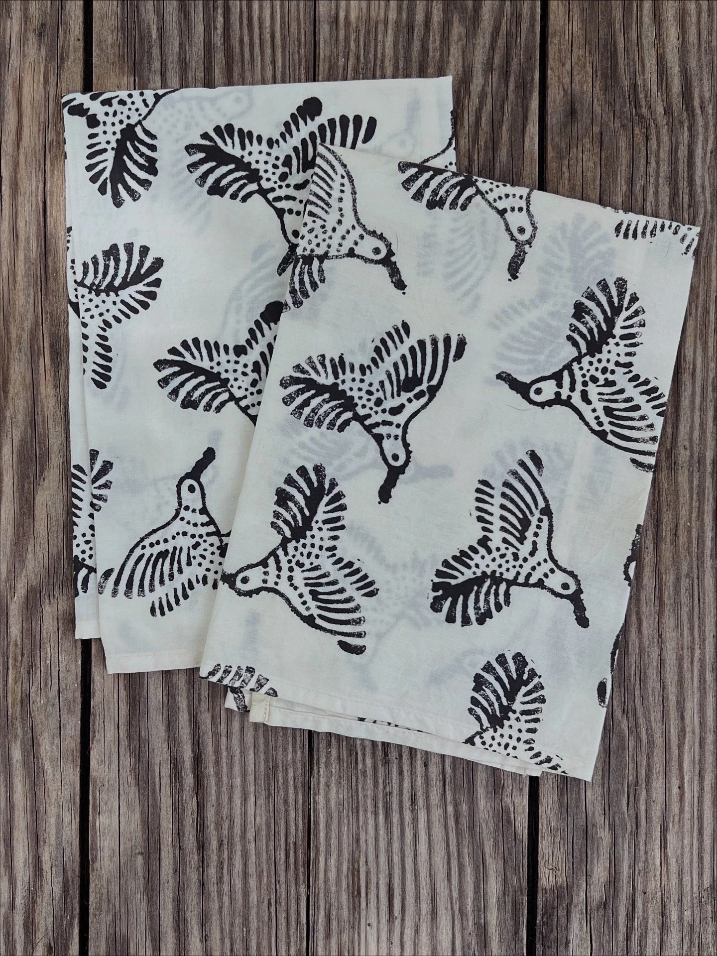 Stack of white cloth napkins with blackbird and feather pattern, placed on a wooden surface.