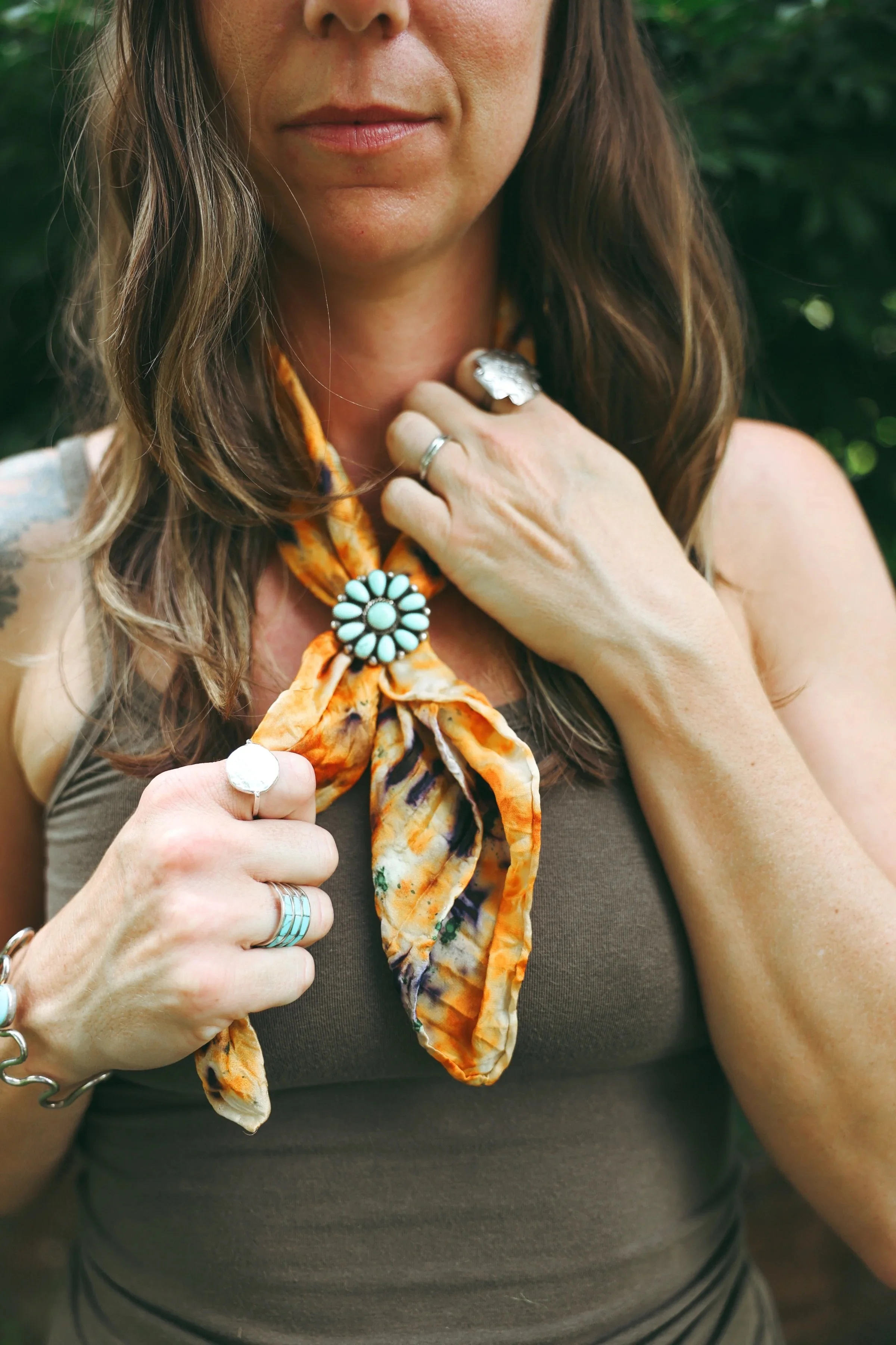 A woman with long brown hair wearing a brown tank top is holding a colorful scarf with orange and yellow hues around her neck, outdoors with green foliage in the background. She has multiple rings on her fingers and a silver bracelet on her wrist.