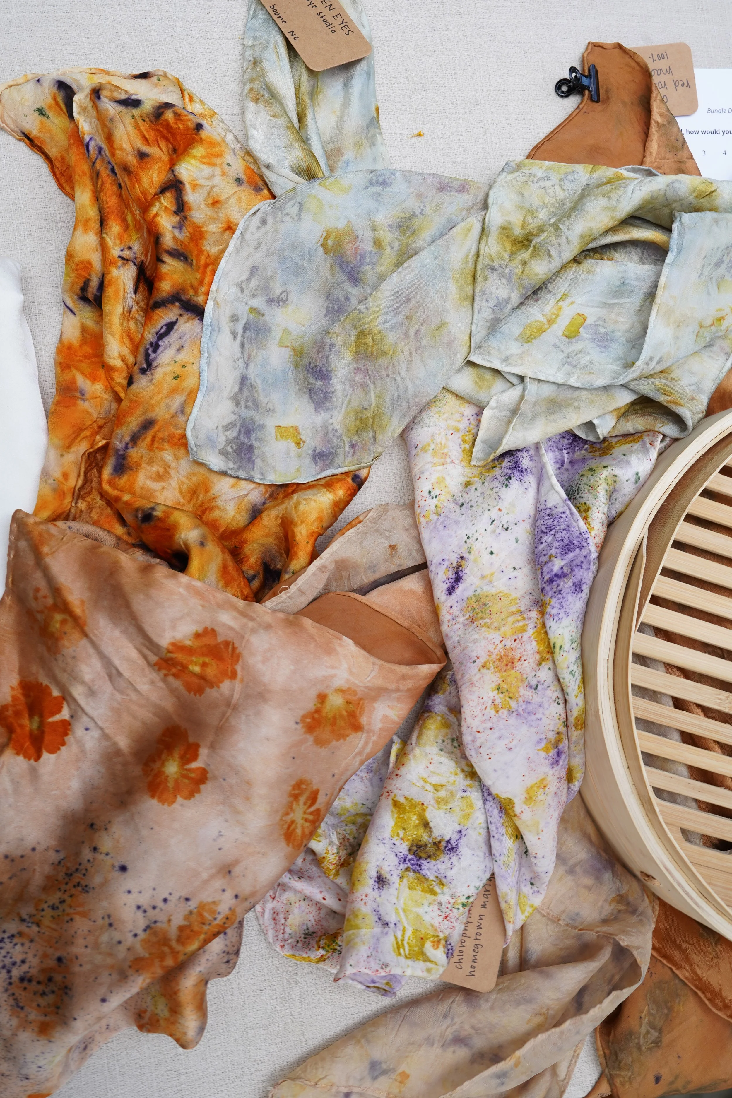 Various crumpled, colorful fabric pieces with paint splatters and watercolor-like patterns, some with tags attached, surrounding a wooden slatted object.