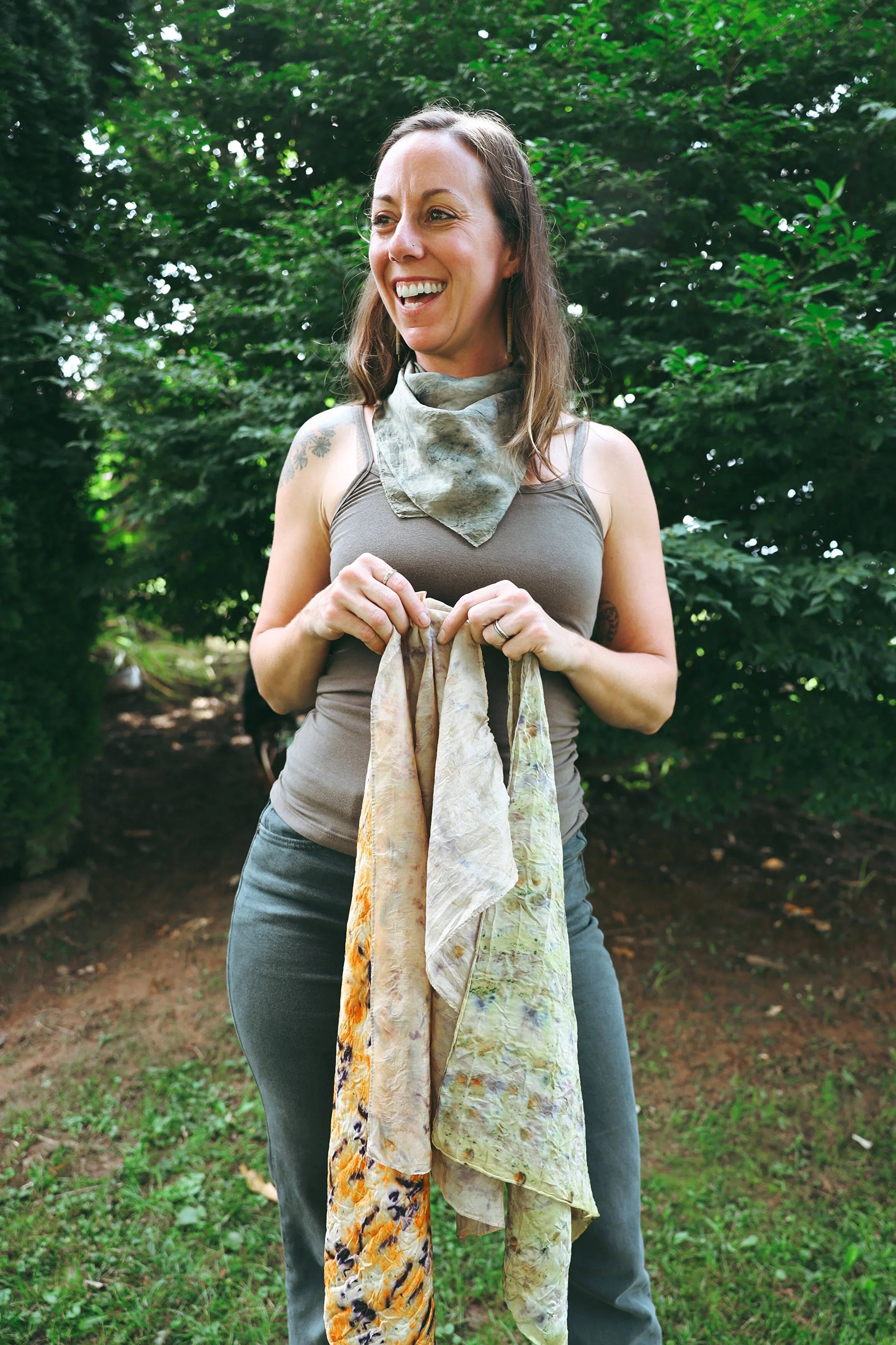 A smiling woman standing outdoors holding several colorful, patterned fabrics or scarves, with green trees and foliage in the background.