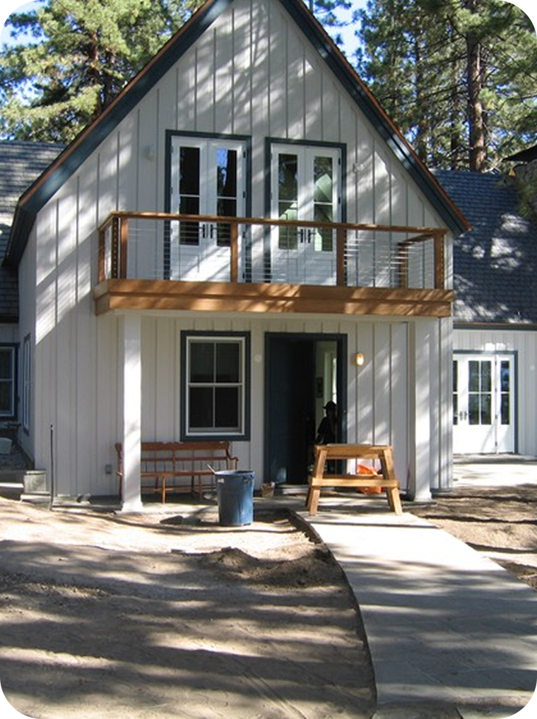 New two-story house with white vertical siding, dark trim, and a wooden balcony on the second floor, situated among tall pine trees.