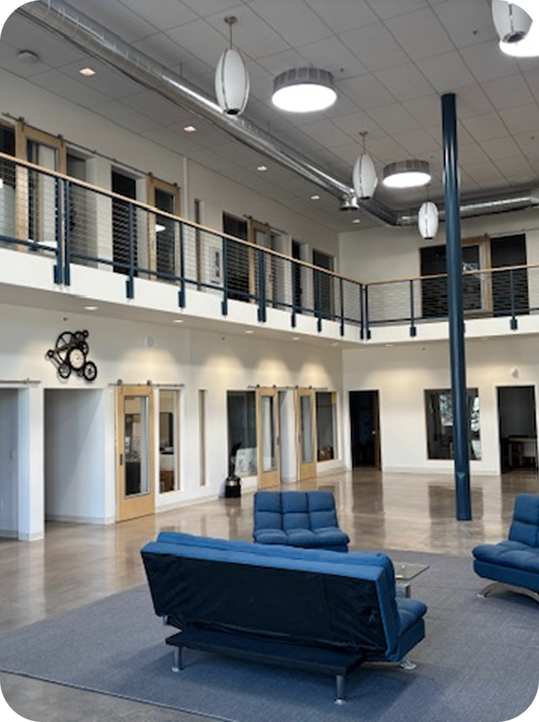 Modern lobby area with blue couches, wooden accents, a high ceiling with hanging white lights, and a second-floor balcony with black railing.
