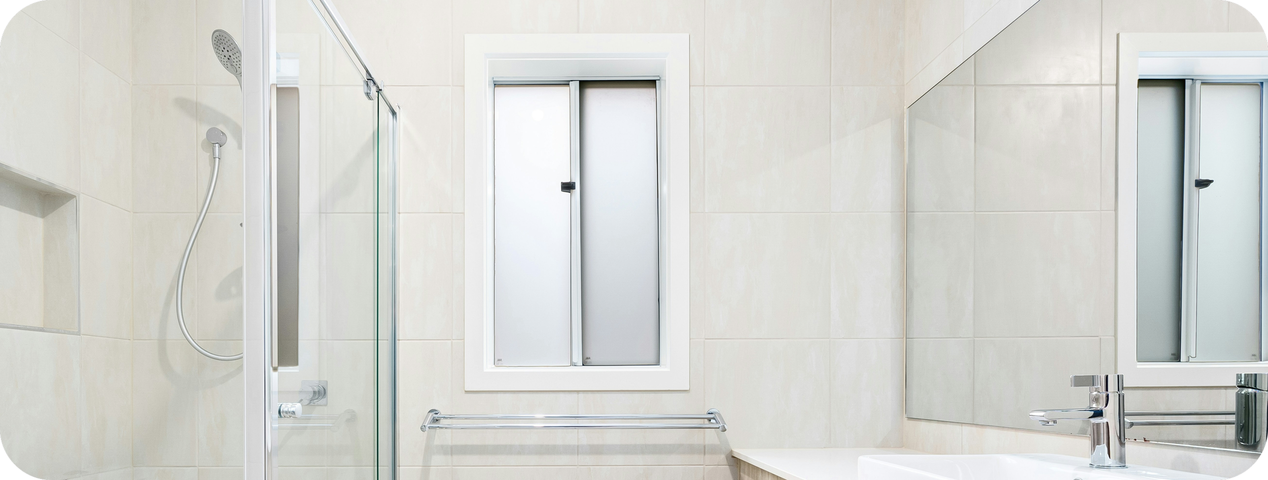 Bathroom with beige tiled walls, a frosted sliding window, a mirror, a white sink with a chrome faucet, a towel bar, and a shower area with a glass door and handheld showerhead.