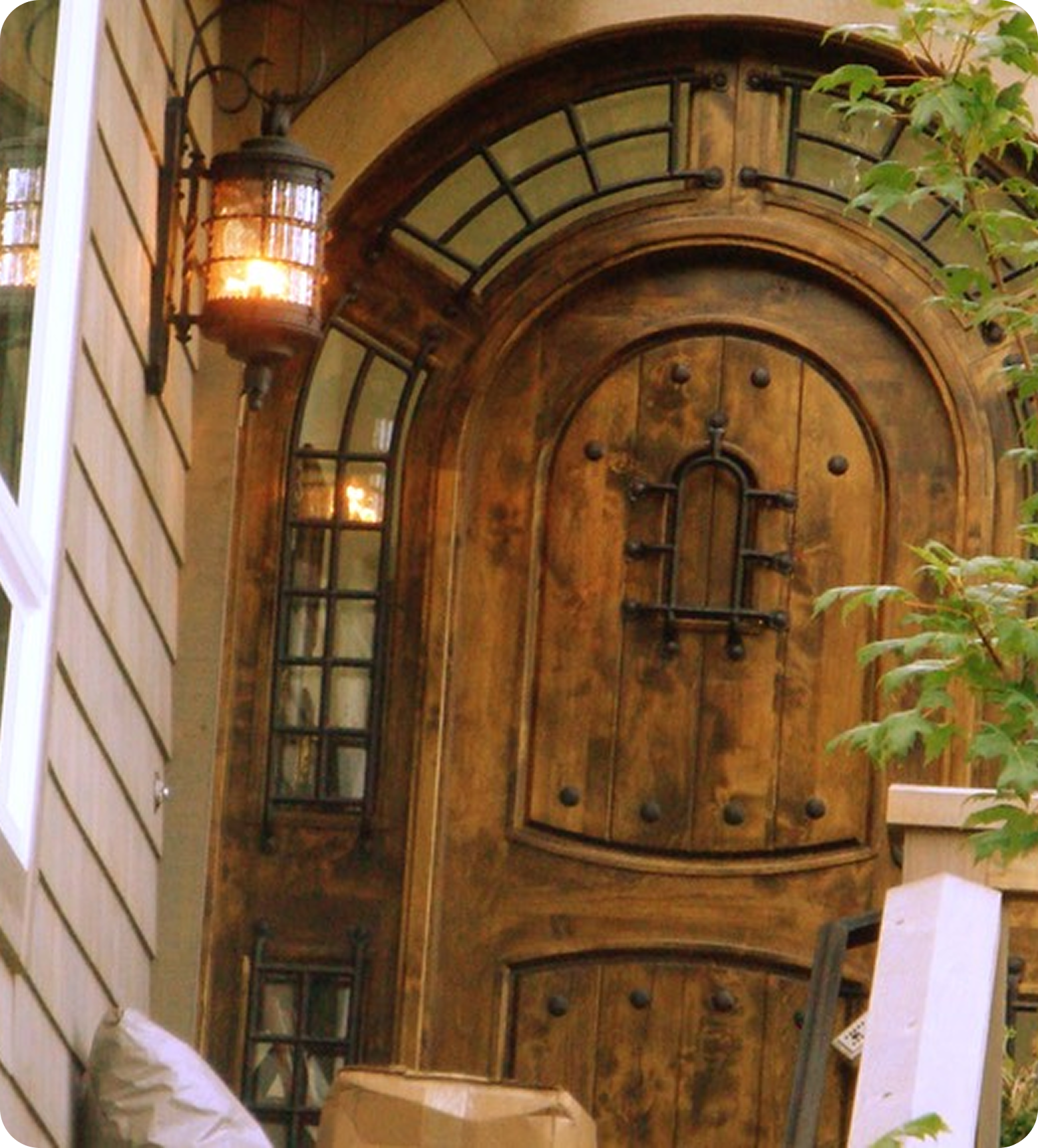 A wooden front door with a rounded top, black iron hardware, and decorative black iron details. To the left, a wall-mounted lantern-style light fixture is lit. There is a small windowed side panel next to the door. Green leafy plants are partially visible on the right side.