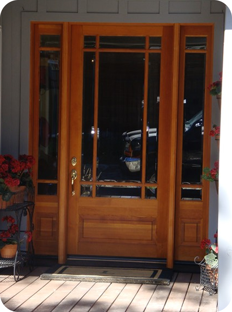 Front door with wooden frame and glass panels, surrounded by potted plants and a doormat, leading to a porch or entrance area.