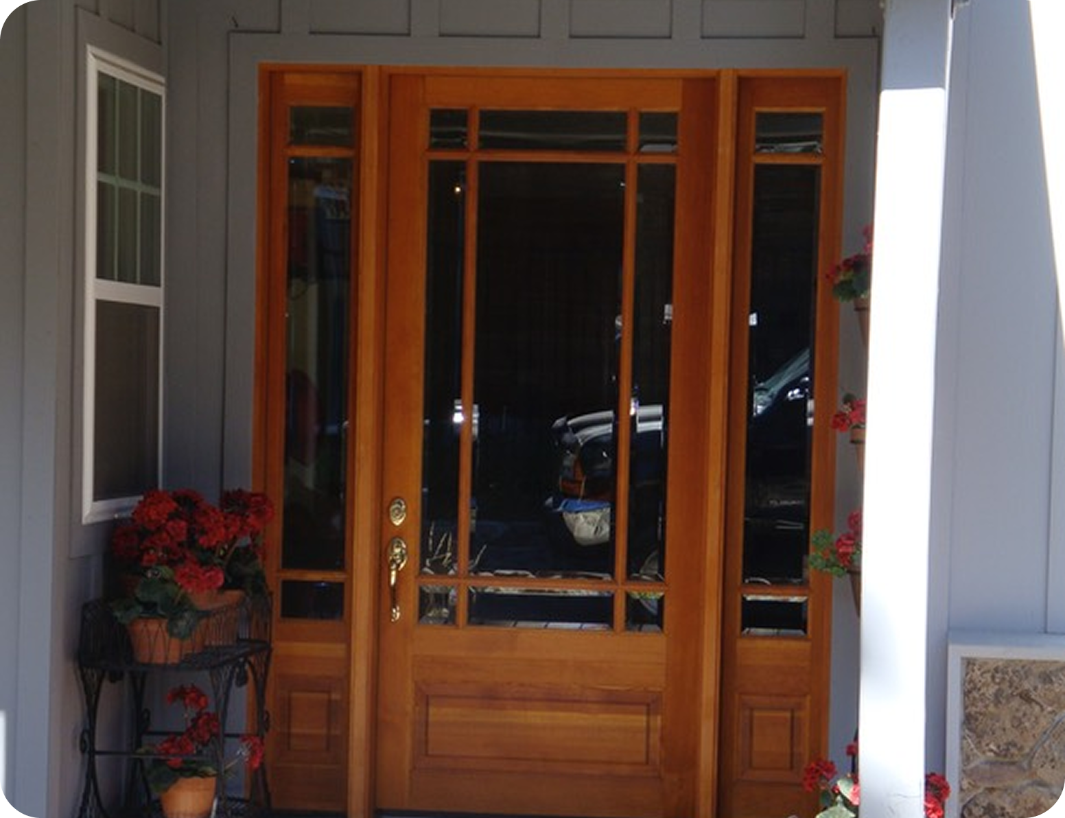 Front door with wood frame and glass panels, flanked by two flower pots with red flowers, with an off-white exterior wall.