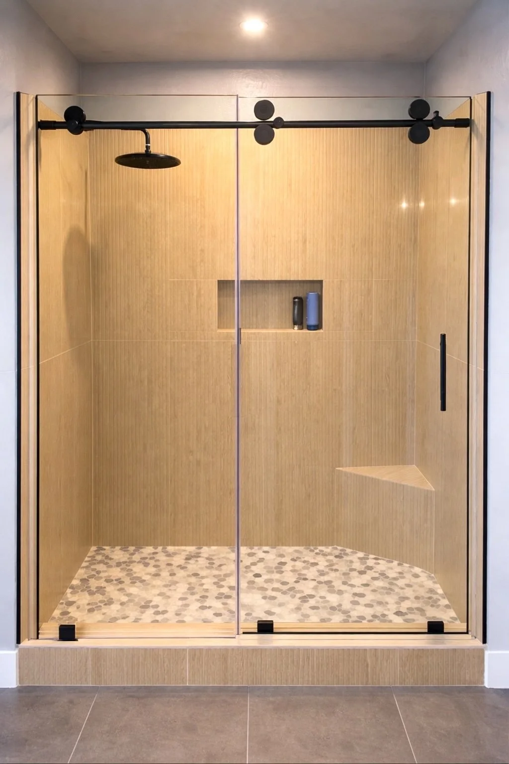Modern walk-in shower with glass doors, black rain showerhead, and wood-paneled walls, including built-in niche with toiletries and a corner seat.