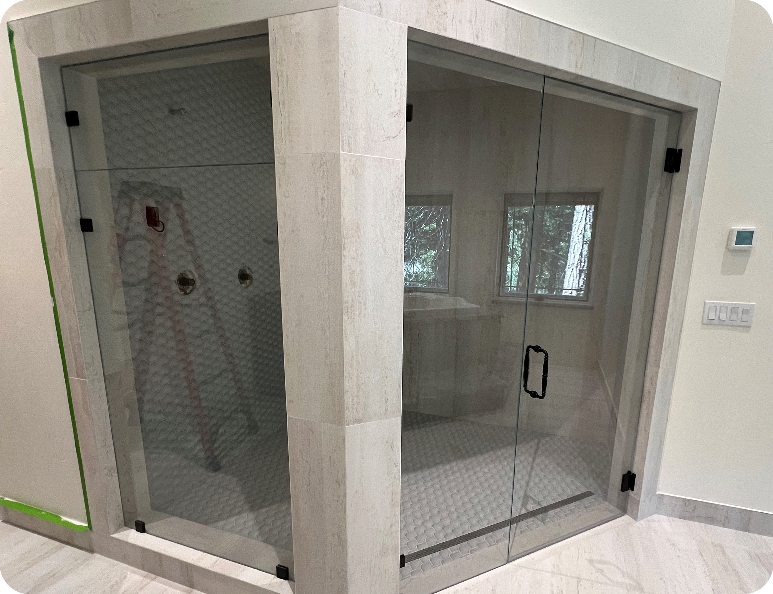 A modern bathroom shower enclosure with glass walls and door, tiled in light-colored stone with a textured pattern, featuring two separate shower sections visible through the glass.