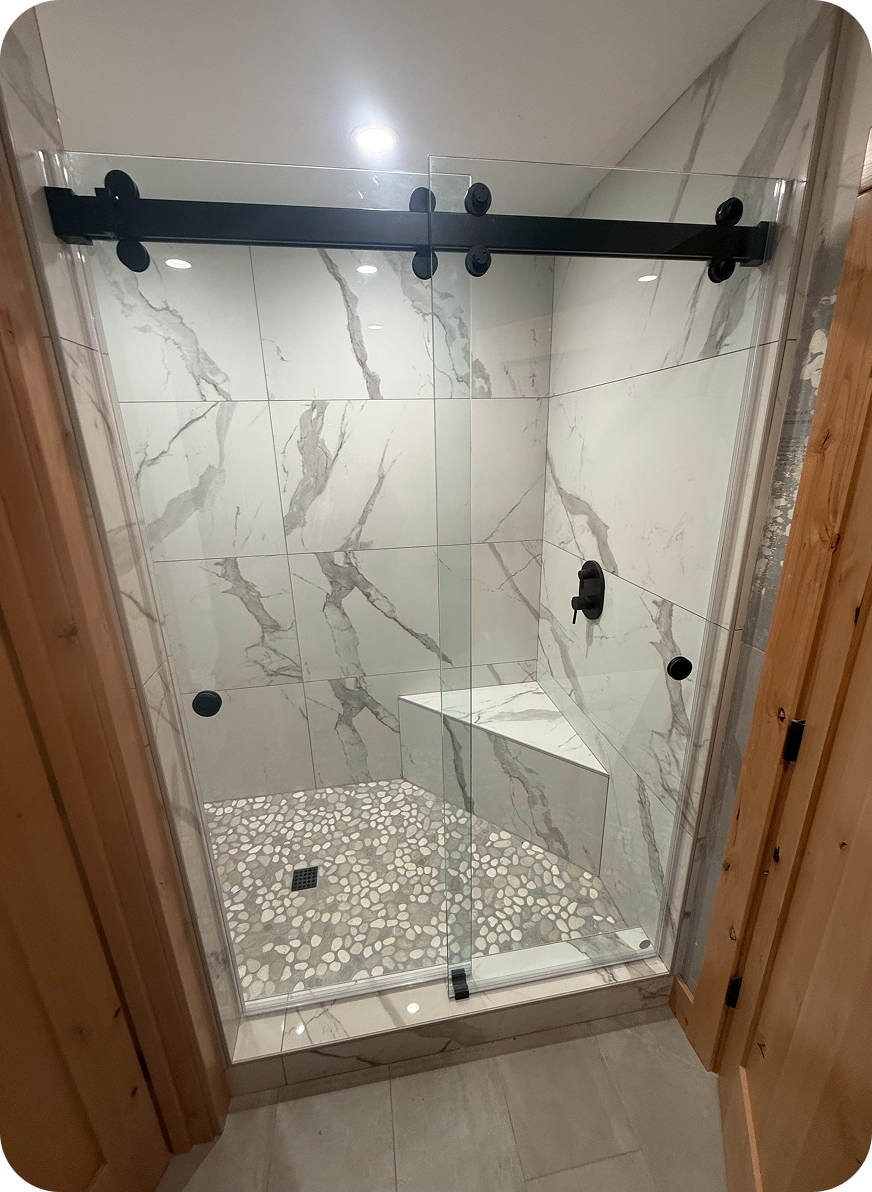 A glass shower enclosure with black hardware, marble tile walls with gray veining, pebble tile floor, and a small built-in marble bench.