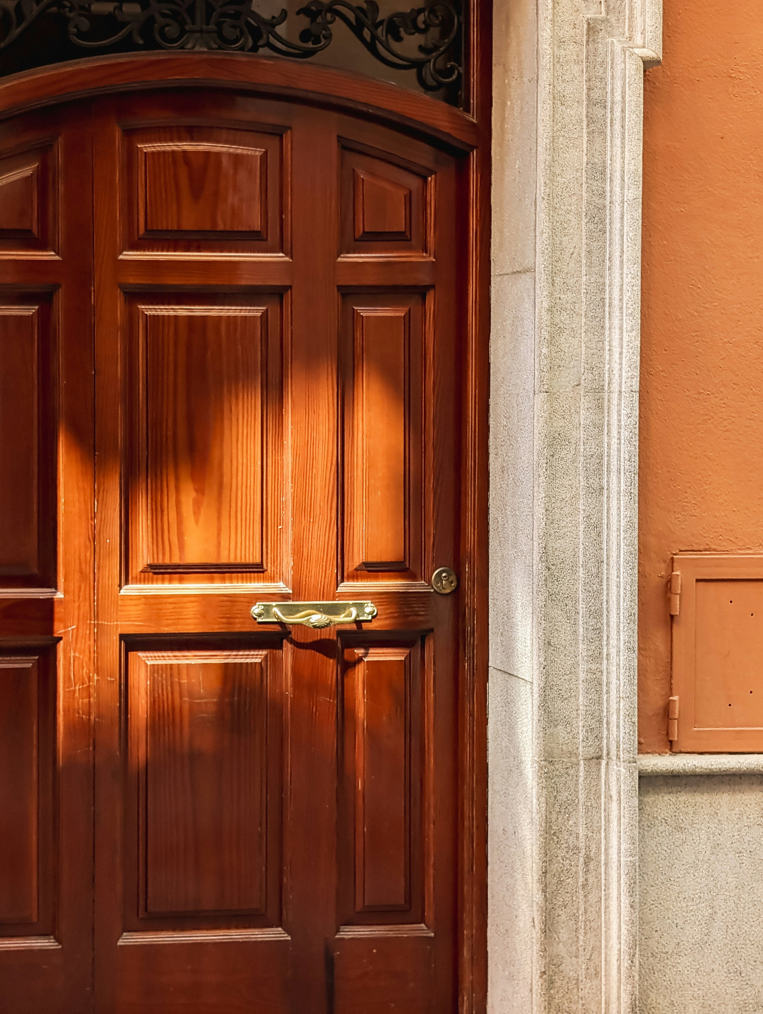 A closed wooden door with rectangular panels and a gold-colored handle, set in a building with stone trim and an orange wall.