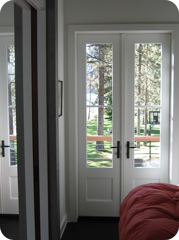 Interior view of a room with glass double doors leading to an outdoor area with trees and a grassy yard. Part of a bed with a red or coral blanket is visible in the lower right corner.