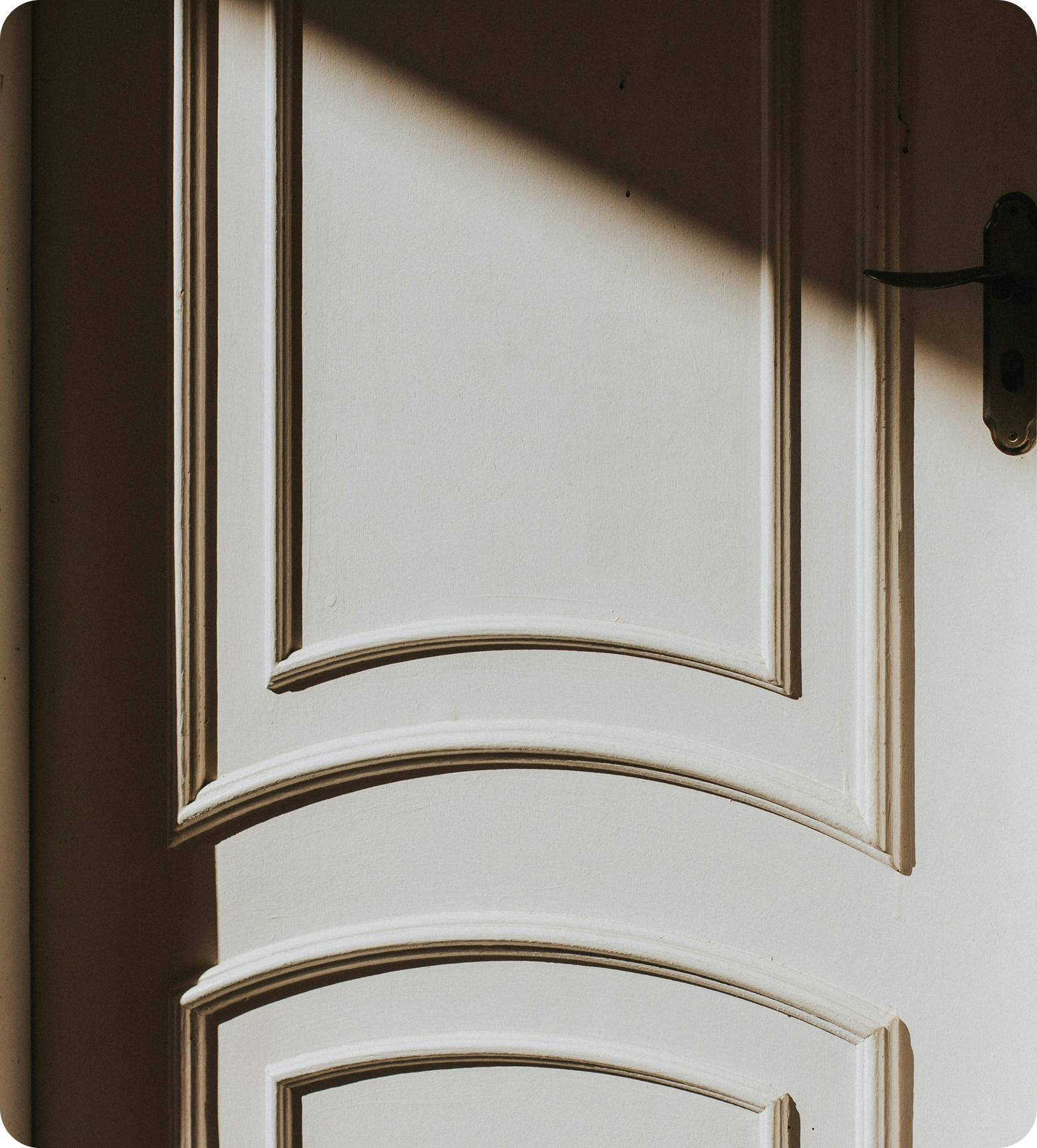 Close-up of a white wooden cabinet door with black hinges and a black handle, partially open, with sunlight casting a shadow on the door and wall.