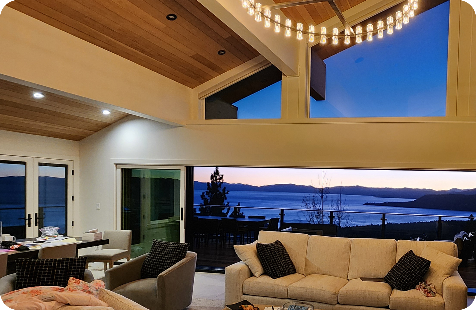 Living room with large windows showing a lake and mountain landscape at sunset, modern furniture, and a chandelier on a wooden ceiling.
