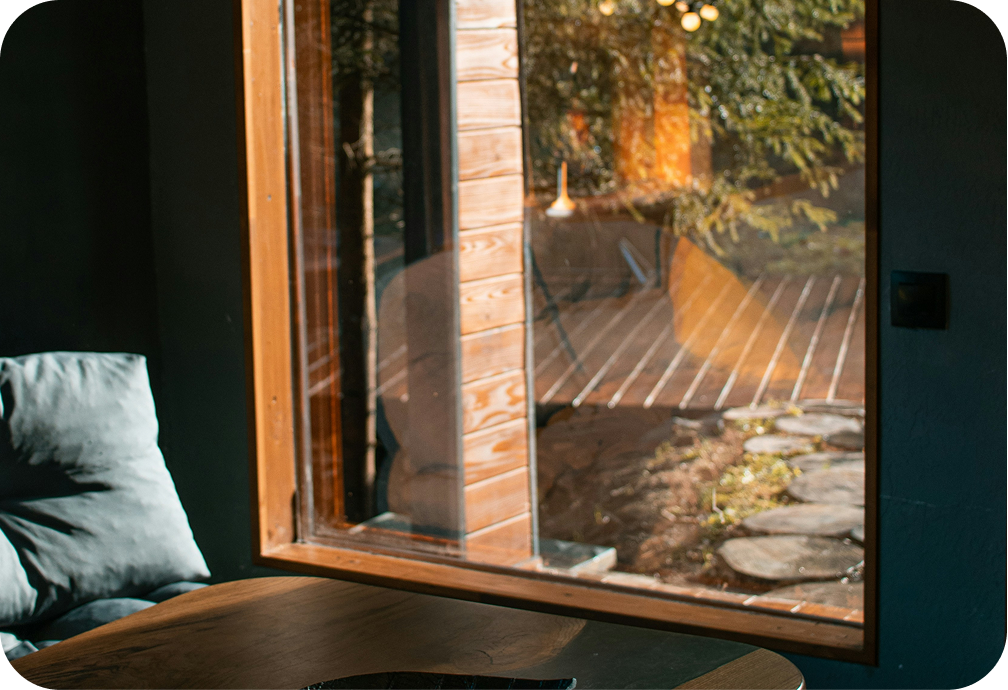 Sunlight shines through a large wooden-framed window, illuminating a cozy interior with a gray cushion on a dark wall in the foreground and an outdoor garden area with a wooden deck, stepping stones, and trees outside.