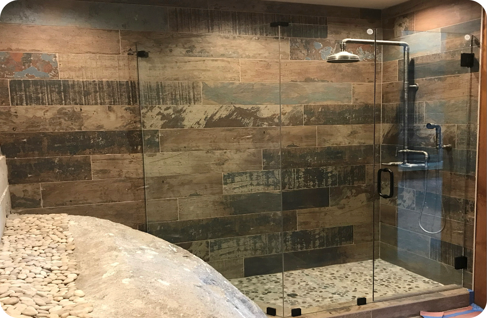 A modern bathroom shower with glass doors, wooden wall tiles, and a pebble stone shower floor.