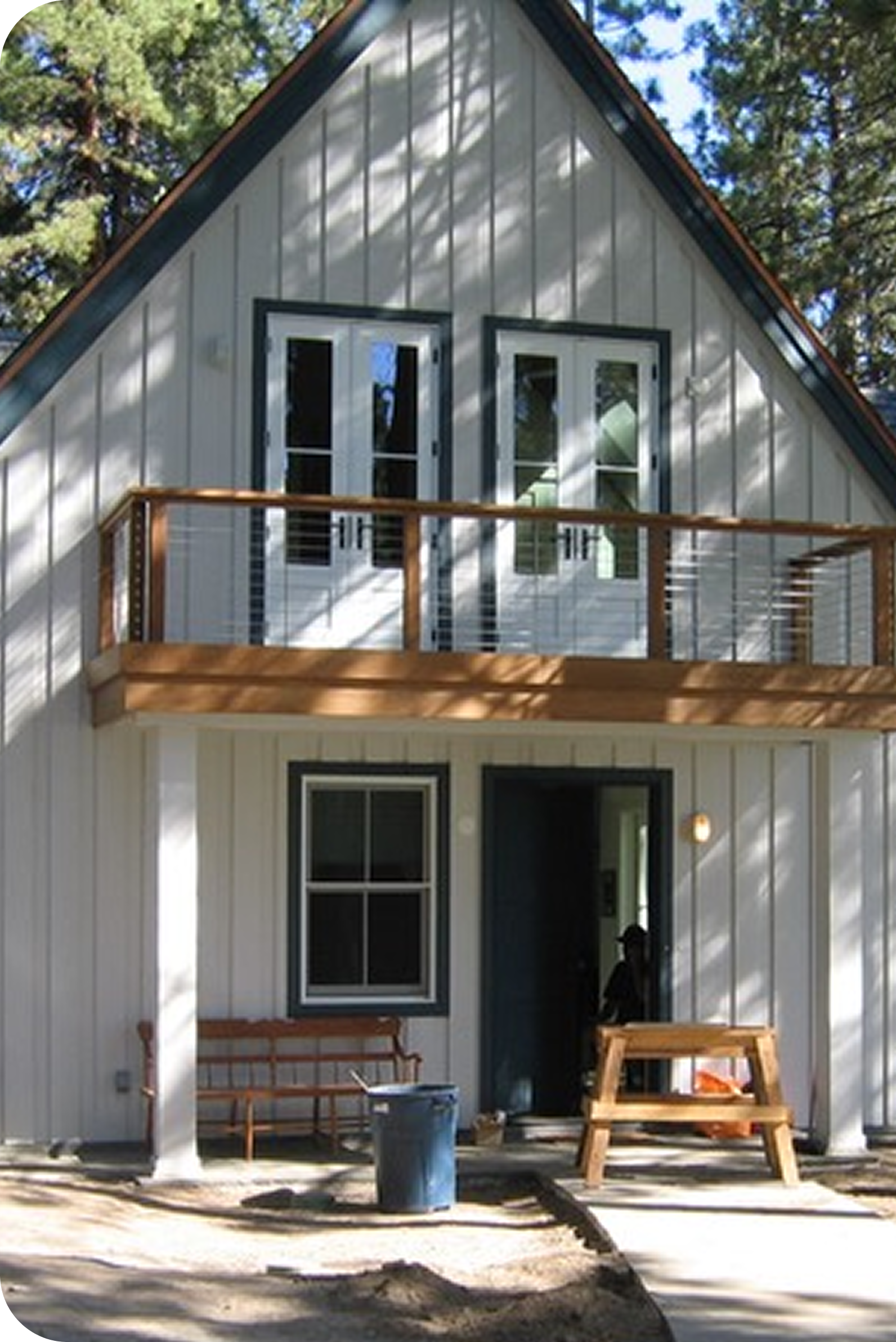 The image shows the exterior of a two-story house with a steep gabled roof. The house has light-colored siding with dark trim around the windows and doors. There is a small balcony with a wooden railing on the upper level. The lower level has a sliding glass door and a window. There are outdoor furniture pieces including a wooden bench and a small table, and a trash can in front of the house. The area around the house appears to be under construction or renovation, with visible dirt and some construction materials.