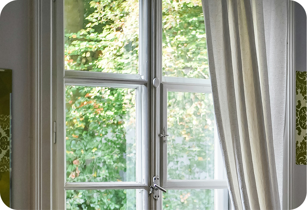 A white window with four panes, partially covered by a cream-colored curtain, revealing a view of green leafy trees outside.