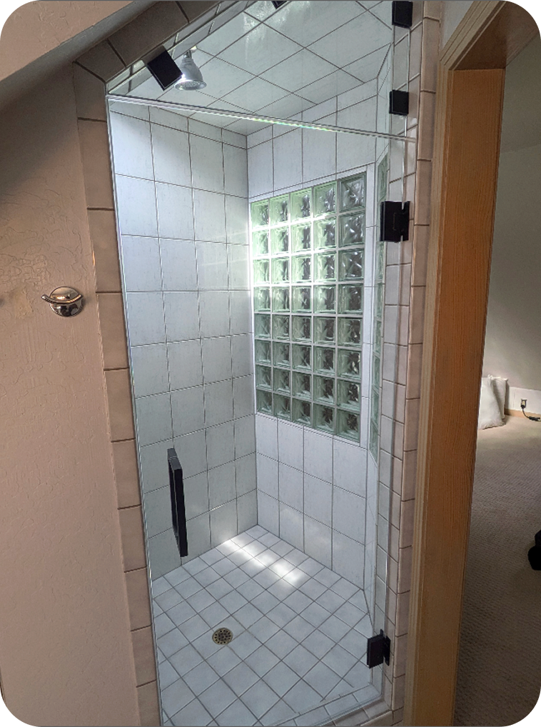 A small glass-enclosed shower with tiled walls and floor, a glass block window, a ceiling-mounted showerhead, and a drain on the floor, located next to a bathroom doorway.