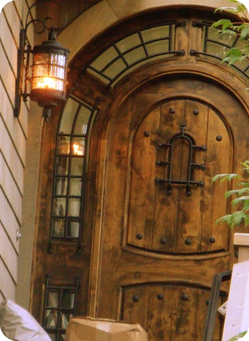 A wooden door with iron hardware and an arched window above it, surrounded by wooden framing, with a lantern on the left and some plants on the right.