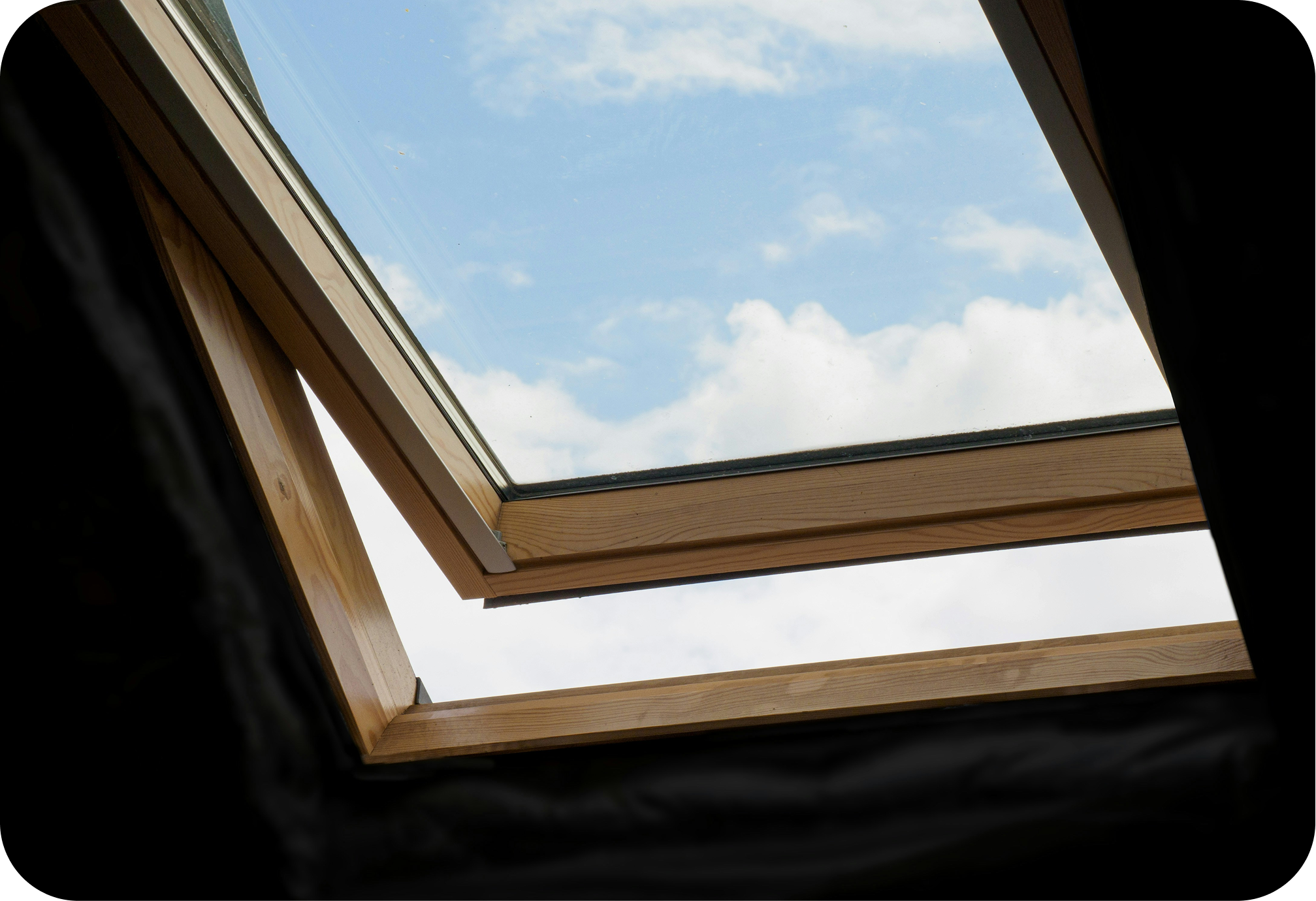 View of a blue sky with clouds seen through a roof window with wooden framing, from inside a room.